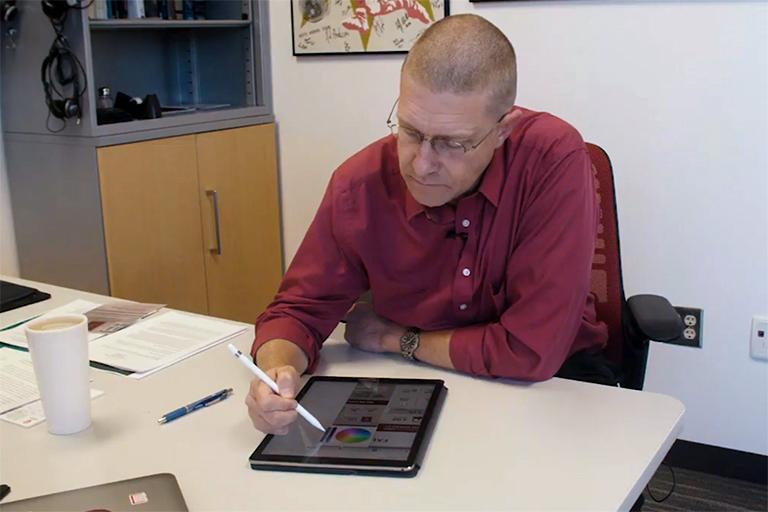 Chris Foley sitting at a desk while grading student work using an Apple Pencil and an iPad.
