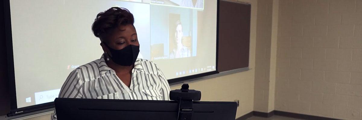 Photo of an instructor at the teaching computer with students participating via Zoom behind her
